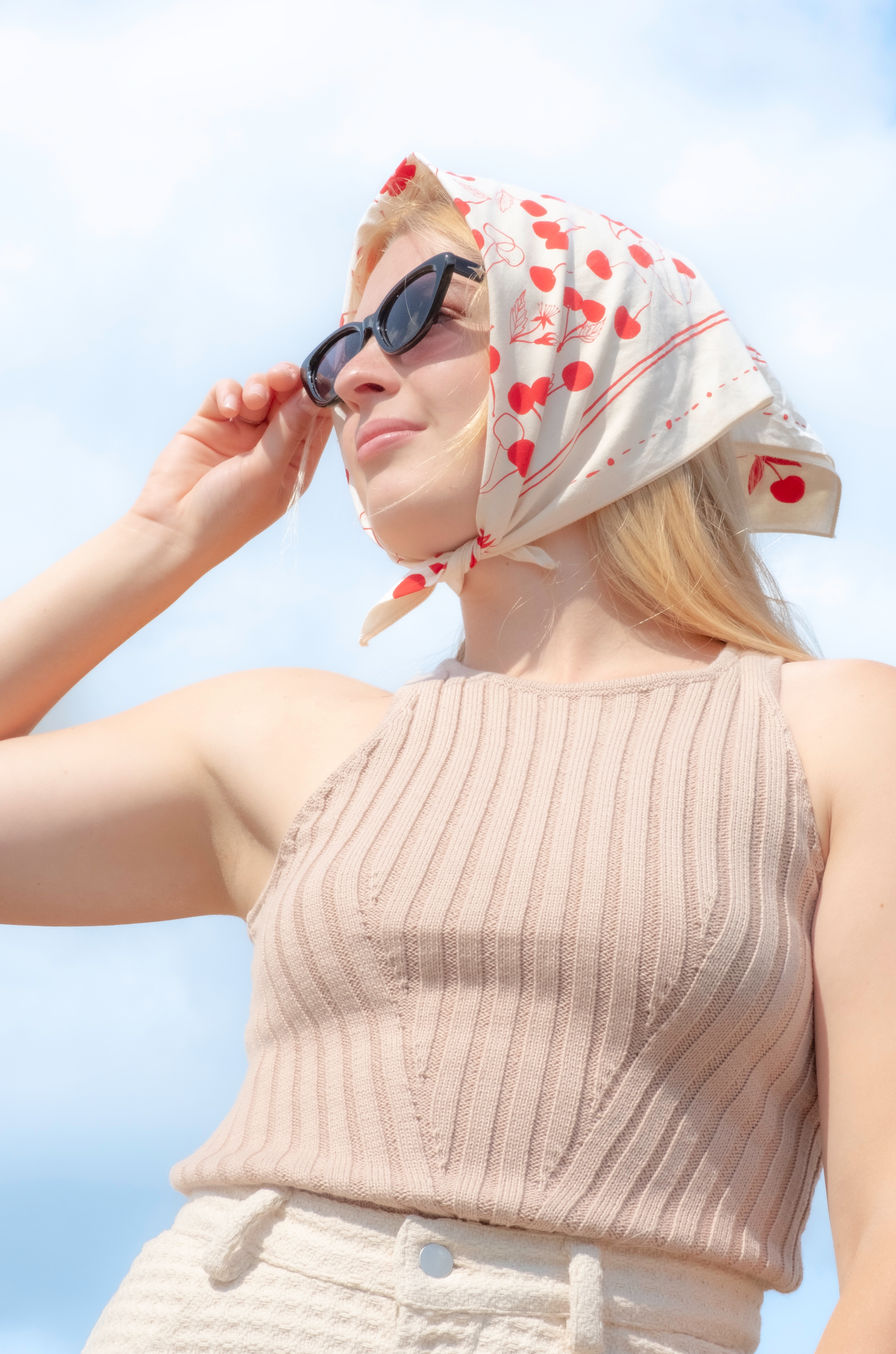 Woman wearing a beige sleeveless top and white pants with a red and white patterned headscarf against a clear blue sky.